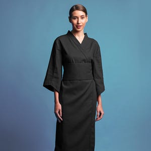 May include: A black kimono-style wrap dress with a matching belt. The dress has wide sleeves and a V-neckline. The model is wearing black sneakers. The background is a gradient of blue.