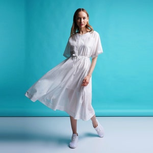 May include: A woman wearing a white, flowing dress with a cinched waist and short sleeves. She is standing on a light blue background and wearing white sneakers.