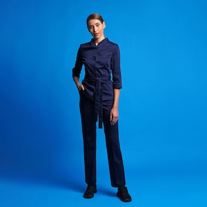May include: A woman wearing a dark blue uniform with a belt and long sleeves. She is standing in front of a blue background.