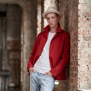 May include: A red jacket with a white t-shirt and light blue jeans. The jacket has a button-up front and a collar. A beige fedora hat completes the look. The person is leaning against a brick wall.