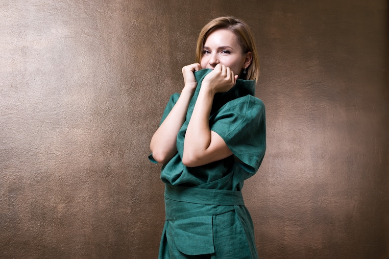 May include: A woman wearing a teal green linen dress with a high neckline, standing in front of a brown background. She is smiling and holding the collar of her dress with both hands.