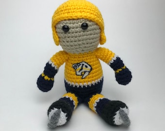 May include: A crocheted plush toy of a hockey player wearing a yellow and blue jersey with a team logo. The player is wearing a helmet and skates.