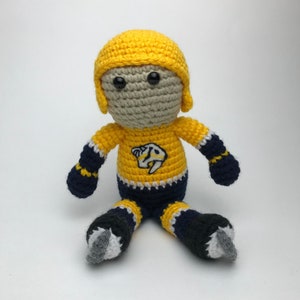 May include: A crocheted plush toy of a hockey player wearing a yellow and blue jersey with a team logo. The player is wearing a helmet and skates.