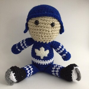 Peut inclure: Poupée de joueur de hockey en crochet portant un chandail bleu et blanc avec une feuille d'érable blanche. La poupée porte un casque bleu et des patins noirs.