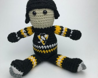 May include: A crocheted plush toy of a hockey player wearing a black and yellow jersey with a penguin logo. The toy has a black helmet, black pants, and white and yellow stripes on the sleeves and legs.
