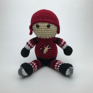 May include: A crocheted plush toy of a hockey player wearing a red, white, and black uniform with a team logo. The toy is sitting on a white surface.