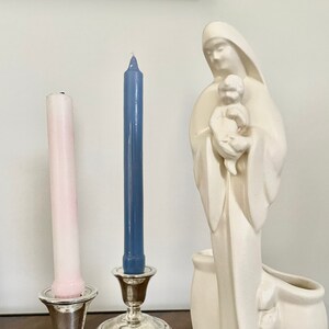 Op de afbeelding: Een wit keramiek beeld van de Maagd Maria die een baby Jezus vasthoudt. Het beeld heeft de vorm van een vaas met een decoratief bloemenontwerp. Op de achtergrond staan twee zilveren kandelaars met roze en blauwe kaarsen.