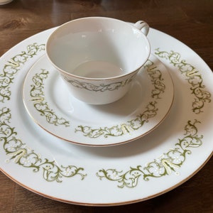 Puede incluir: Un juego de taza y platillo de té de porcelana blanca con un diseño floral verde y dorado. La taza tiene un asa y el platillo tiene un borde festoneado.