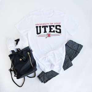 May include: White t-shirt with the text "University of Utah" and "UTES" in black. The logo features a red "U" with a feather design.
