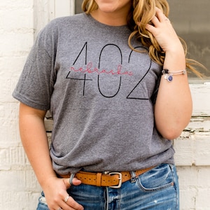 May include: A gray t-shirt with a black outline of the number 402 and the word "Nebraska" written in red cursive inside the outline.