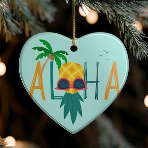 Pode incluir: Um ornamento de cerâmica em forma de coração com fundo azul claro, apresentando um abacaxi amarelo usando óculos de sol vermelhos. A palavra ALOHA está impressa em letras laranja e verde-escuras. Uma palmeira verde e pássaros são visíveis.