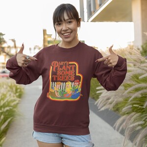 以下が含まれることがあります： 「Let's Plant Some Trees」というレトロなデザインのグラフィックがプリントされた、マルーンのトレーナーを着た女性。トレーナーには、木を植えている子供たちの漫画が描かれている。彼女は両手でトレーナーを指差している。