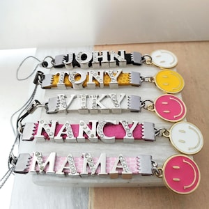 Könnte beinhalten: Nahaufnahme von fünf personalisierten Armbändern. Jedes Armband zeigt einen Namen, der in silbernen Buchstaben mit klaren Strasssteinen geschrieben ist. Die Bänder sind in verschiedenen Farben, darunter Schwarz, Gelb, Rosa und ein rosa gemustertes Design. Jedes Armband hat einen Smiley-Anhänger.
