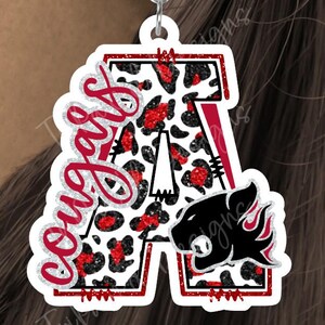 May include: A dangle earring featuring a large letter A with a white, black, and red glitter leopard print pattern. The word Cougars is written in red script across the letter, which also displays a black cougar head logo with red flames.