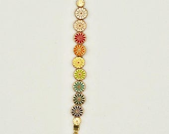 Könnte beinhalten: Eine goldene Kette mit einem Regenbogen aus emaillierten Blumenanhängern. Die Kette hat oben einen Karabinerverschluss und unten einen einzelnen weißen Blumenanhänger.