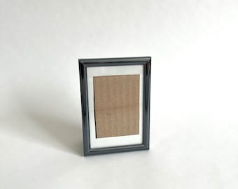 May include: A small, rectangular black picture frame with a white border and a brown cardboard backing. The frame is standing upright on a white surface. The frame is empty.