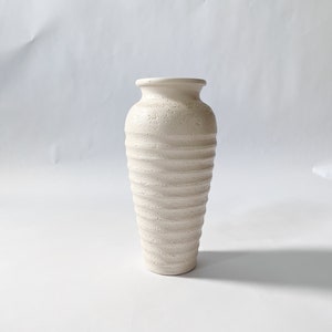 Op de afbeelding: Een witte keramische vaas met een getextureerd, geribbeld ontwerp. De vaas is hoog en slank, met een ronde basis en een licht uitlopende opening.