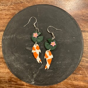 Peut inclure: Une paire de boucles d'oreilles pendantes avec un nénuphar vert et rose et un charme de poisson-koi blanc et orange suspendu en dessous.