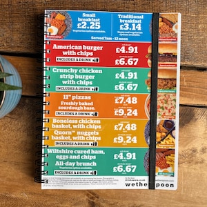 Puede incluir: Un menú colorido con una banda elástica negra. El menú enumera opciones de desayuno y almuerzo, que incluyen desayuno pequeño, desayuno tradicional, hamburguesa americana con patatas fritas, hamburguesa de pollo crujiente con patatas fritas, pizzas de 28 cm, cesta de pollo deshuesado con patatas fritas, cesta de nuggets de Quorn con patatas fritas y jamón curado de Wiltshire, huevos y patatas fritas. El menú también incluye los precios de cada artículo.