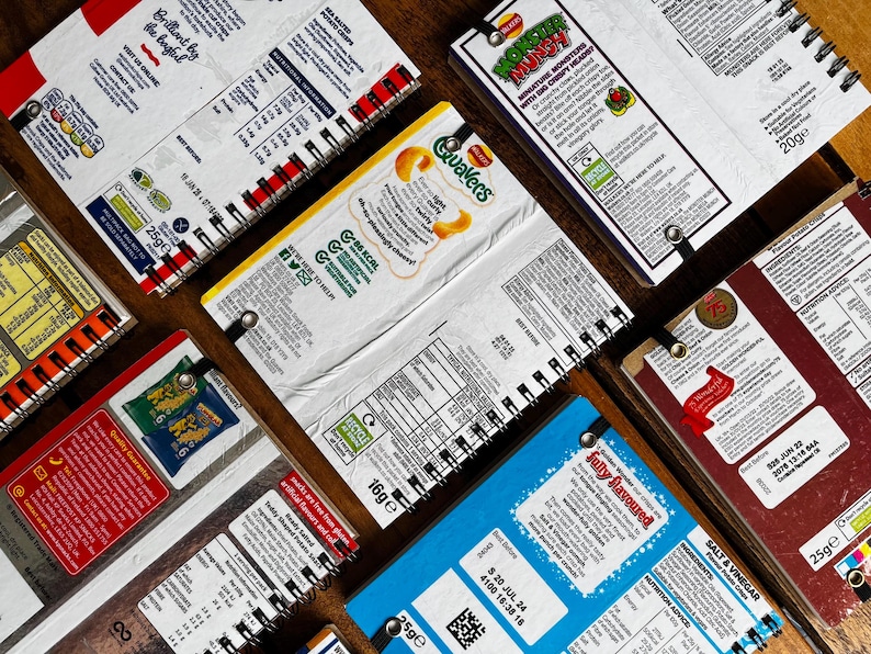 Puede incluir: Una colecci&oacute;n de ocho peque&ntilde;os cuadernos de espiral con diferentes cubiertas de envases de marcas de alimentos. Las cubiertas presentan marcas como Quavers, Monster Munch y Salt & Vinegar. Los cuadernos est&aacute;n todos cerrados y dispuestos en semic&iacute;rculo sobre una superficie de madera.
