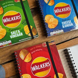 Puede incluir: Tres cuadernos diferentes de patatas fritas Walkers. Los cuadernos son rojo, verde y azul. Cada cuaderno tiene un sabor diferente de patatas fritas Walkers en la parte delantera. El cuaderno rojo tiene el texto "Some see potatoes, we see potential" y "0% Great British Potato" en la parte delantera. El cuaderno verde tiene el texto "Seriously Salt & Vinegar" en la parte delantera. El cuaderno azul tiene el texto "Glorious Cheese & Onion" en la parte delantera.
