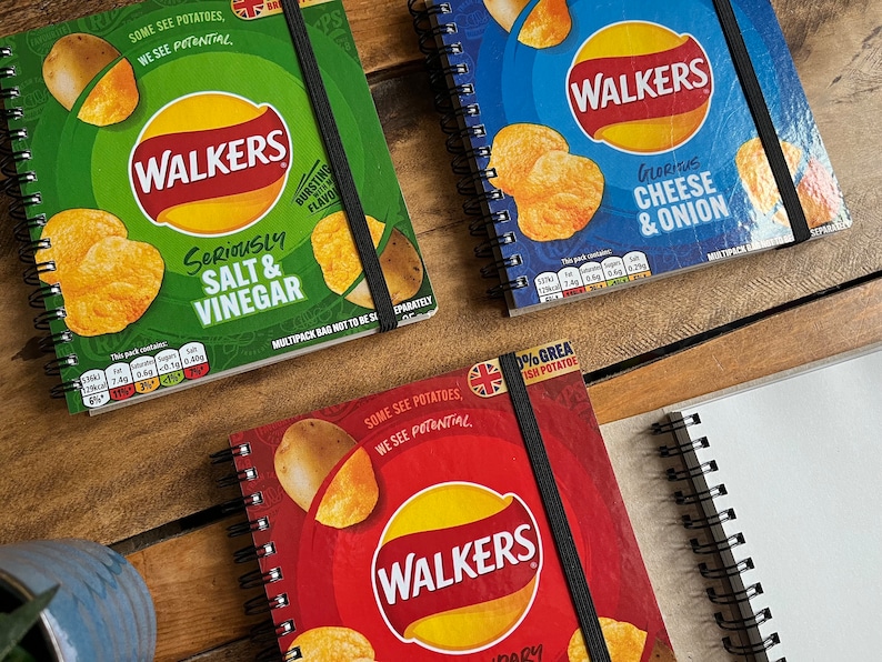 Puede incluir: Tres cuadernos diferentes de patatas fritas Walkers. Los cuadernos son rojo, verde y azul. Cada cuaderno tiene un sabor diferente de patatas fritas Walkers en la parte delantera. El cuaderno rojo tiene el texto "Some see potatoes, we see potential" y "0% Great British Potato" en la parte delantera. El cuaderno verde tiene el texto "Seriously Salt & Vinegar" en la parte delantera. El cuaderno azul tiene el texto "Glorious Cheese & Onion" en la parte delantera.