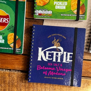 Puede incluir: Una libreta azul con cierre de banda el&aacute;stica negra. La portada presenta una ilustraci&oacute;n blanca de una persona haciendo patatas fritas con el texto "KETTLE SEA SALT & Balsamic Vinegar of Modena ABSOLUTELY NOTHING ARTIFICIAL".