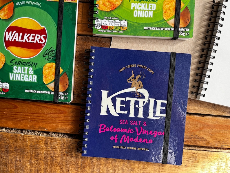 Puede incluir: Una libreta azul con cierre de banda el&aacute;stica negra. La portada presenta una ilustraci&oacute;n blanca de una persona haciendo patatas fritas con el texto "KETTLE SEA SALT & Balsamic Vinegar of Modena ABSOLUTELY NOTHING ARTIFICIAL".