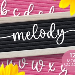 May include: A black felt letter board with the name "melody" in white cursive letters. Additional white cursive letters are scattered around the board. The text "12 MORE COLORS!" is in a pink circle. The background is pink.