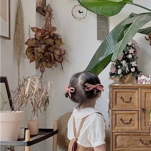 May include: A young girl wearing a white shirt and brown overalls with a bow in her hair. She is standing in a room with a wooden dresser and a plant in the background.