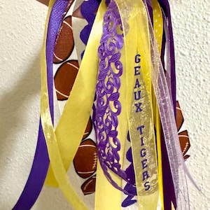 May include: A decorative ribbon cluster featuring purple, yellow, and white ribbons. Some ribbons have a brown football pattern, while one yellow ribbon displays the purple text GEAUX TIGERS. A purple lace ribbon is included in the arrangement.