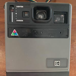 Peut inclure: Un appareil photo instantané Kodak vintage gris et noir, avec un objectif, un viseur et des commandes pour éclaircir et assombrir les images. L'appareil photo a une sangle multicolore et le logo Kodak.