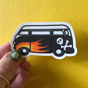 May include: A die-cut sticker featuring a black van with a white roof. The side of the van displays a graphic of orange and yellow flames near the front wheel and a white skull and crossbones symbol positioned above the rear wheel.