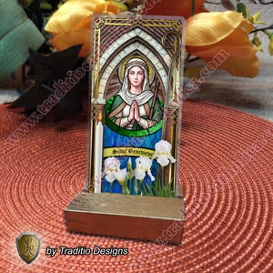 Può includere: Un'opera d'arte religiosa raffigurante Santa Genoveffa, esposta su un supporto di legno. L'opera raffigura una donna in abito verde e bianco, con un'aureola, in preghiera. Sono presenti anche una candela e iris bianchi. Il testo "Saint Genevieve" è visibile.
