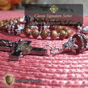 Puede incluir: Primer plano de un rosario con una cruz plateada y cuentas de madera de olivo. La cruz tiene un emblema dorado y está conectada a las cuentas por una cadena plateada. El texto "Classic Signature Series" y "18K GOLD & BETHLEHEM OLIVE WOOD" son visibles.
