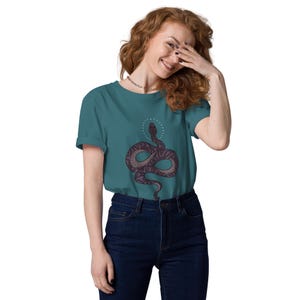 Puede incluir: Camiseta de color turquesa con una ilustración detallada de una serpiente y un halo. La serpiente es de color púrpura oscuro con patrones púrpuras más claros. La camiseta se combina con vaqueros azul oscuro. Estilo casual y artístico.