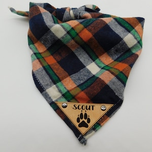 May include: A blue, orange, green and white plaid bandana with a brown leather patch that says "SCOUT" and has a paw print.
