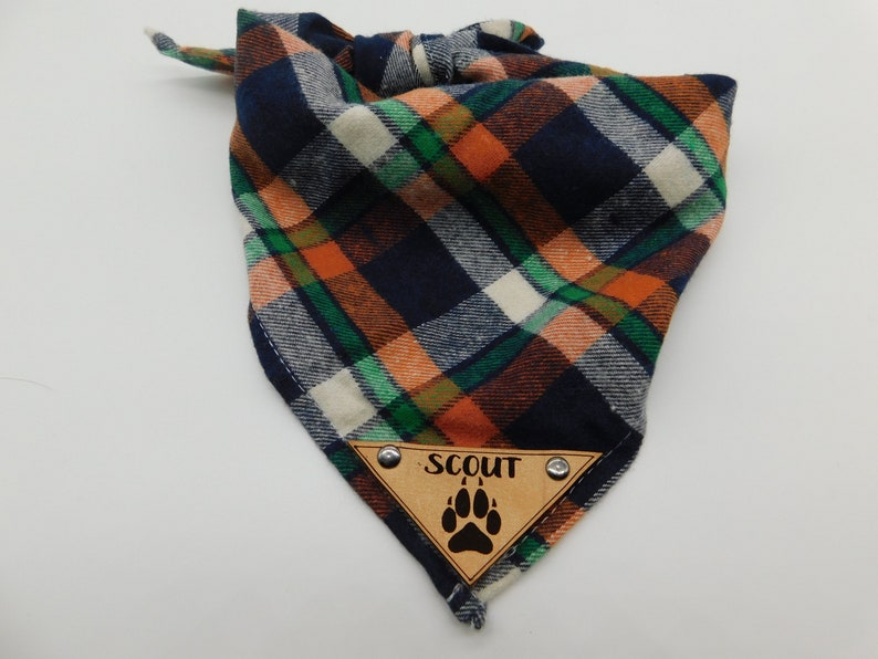 May include: A blue, orange, green and white plaid bandana with a brown leather patch that says "SCOUT" and has a paw print.