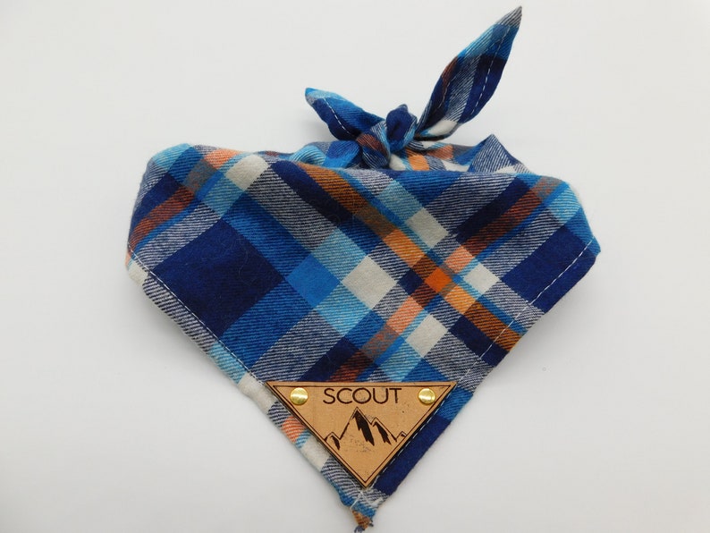 May include: Blue, orange and white plaid bandana with a brown leather tag that says "SCOUT" with a mountain design.
