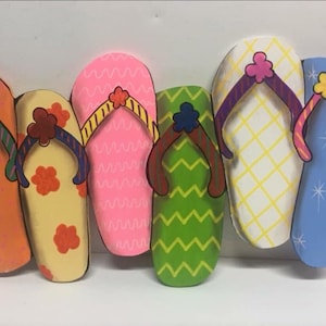 May include: Six colorful wooden flip-flop shaped wall decorations. The flip-flops are painted in various patterns and colors, including orange, yellow, pink, green, white, and blue.