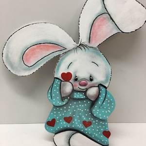 Op de afbeelding: Een wit konijn met roze oren en een turquoise jurk met witte stippen en rode harten. Het konijn houdt zijn handen voor zijn gezicht en heeft een rood hart op zijn wang.