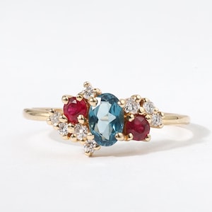 May include: A gold ring with an oval, light blue gemstone at its centre, with two round, red gemstones either side. Small, clear gemstones are clustered around the larger stones. The ring is set against a white background.