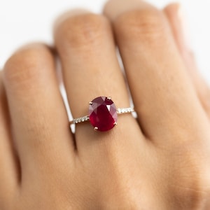 May include: A ring with a large, oval-shaped ruby gemstone set in a white gold band. The ruby is a deep red color and is surrounded by smaller diamonds.