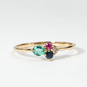 Op de afbeelding: Een gouden ring met een cluster van gekleurde edelstenen. De ring heeft een groene smaragd, een rode robijn, een blauwe saffier en een kleine witte diamant.