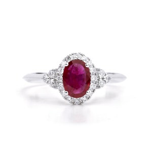 Può includere: Un anello d'argento con un grande rubino ovale circondato da un'aureola di diamanti più piccoli. Il rubino è di colore rosso intenso e i diamanti sono trasparenti.