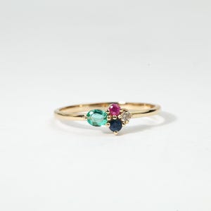 May include: A gold ring featuring a cluster of colorful gemstones. The ring has a vibrant green oval-cut stone, a round magenta stone, a dark blue stone, and a small diamond. The ring is set on a thin gold band.