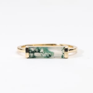 May include: A gold ring with a rectangular, clear gemstone containing green inclusions. The ring has a thin, gold band and gold accents. The gemstone is likely moss agate. The ring is set against a white background.