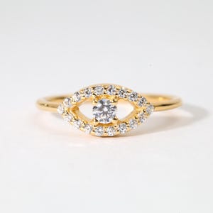 以下が含まれることがあります： 金色のリングで、目の形をしたデザインです。目の中心には、1つの丸い透明な宝石があり、その周りを小さな透明な宝石が囲んでいます。リングのバンドは細く、金色です。