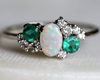 Könnte beinhalten: Ein Ring mit einem zentralen ovalen Opal mit irisierenden Farbreflexen, flankiert von zwei runden grünen Smaragden und akzentuiert durch kleine, klare, rund geschliffene weiße Steine, gefasst in eine polierte Metallschiene mit filigranem Design.