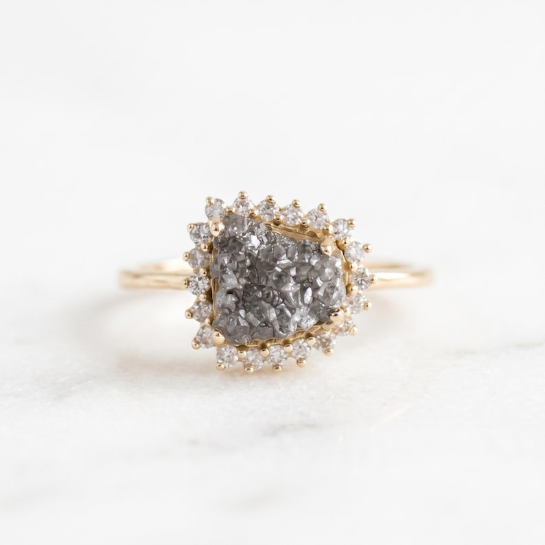 May include: A gold ring featuring a central, irregular, textured gray druzy stone. The stone is surrounded by a halo of small, round, clear white diamonds set in a gold frame. The band of the ring is thin, smooth, and polished gold metal.
